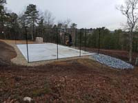 Hillside court primarily for pickleball, accessorized with a basketball goal and fencing, in Plymouth, MA. Side view looks good considering court surface tiles remain to be installed.