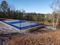 Multicourt for pickleball and basketball, fenced on three sides, on a hillside backyard in Plymouth, MA. Finished blue and grey court with scenic view beyond.
