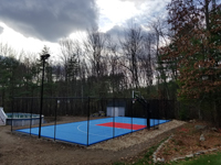 Entire finished blue and red home basketball court in Shirley, MA, oriented at a distance from the back right corner, showing all of the optional fencing and lighting in one image.