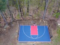 Drone view of royal blue and red backyard basketball court with post to add net games, still in raw surroundings in Sudbury, MA.