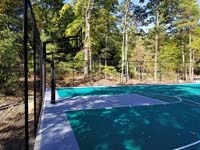 View of hoop end of large emerald green and titanium backyard basketball court in Bolton, MA, with emphasis on the goal system and optional fencing.
