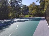 Sun-soaked view of most of length of large emerald green and titanium backyard basketball court in Bolton, MA.