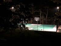 Night time view of large emerald green and titanium backyard basketball court in Bolton, MA, lit by optional LED lighting system for play after hours.