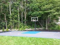 Backyard basketball court with gray and blue sport surface, hoop, fence, and lighting option in West Bridgewater, MA.