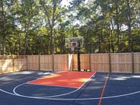 Finished picture of bulk of charcoal and orange home basketball court, looking toward hoop, showing part of custom fence that incorporates traditional cedar wood with rebounder mesh segments, in Walpole, MA.