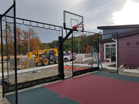 Small slate green and burgundy basketball court at new hotel contruction in Lincoln, RI, highlighting goal and containment netting with construction equipment in future parking lot beyond.