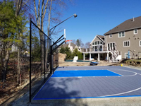 Lengthwise view of most of blue and grey basketball court from left end, showing off backstop fence and optional light system installed above hoop, in Middleton, MA.