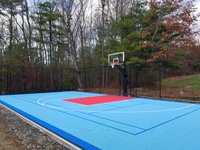 Angled view of most of court from front right, tiled in a surface of three colors: light blue, red, and royal blue. Basketball lines in white and pickleball lines in blue for dual use. Residential basketball in Shirley, MA.
