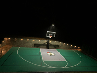Elevated view of entire backyard basketball court from front toward hoop at night, showing optional lighting system in action in Upton, MA.