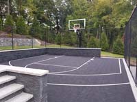 View from entrance through fence, lengthwise toward hoop across black basketball court with extensive associated hardscape work in Weston, MA.