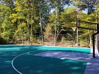 Angled view from corner beside one hoop out at part of large emerald green and titanium backyard basketball court in Bolton, MA.