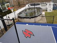 Custom red H logo on small blue and grey backyard basketball court sport surface with accessories in Braintree, MA, adjacent to existing pool.