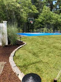 Blue and gray residential basketball court in Easton, MA, viewed from left side of yard, highlighting landscaping by Evergreen.