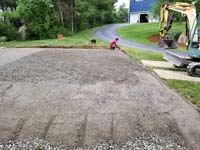 View of packed gravel that precedes the concrete base and the red and grey home basketball court in Groton, MA.