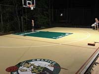 Kids checking out new backyard basketball court under the lights in Londonderry, NH, featuring custom logos and text on a tan and green sport surface