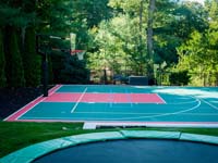 Large green and red (that in some pictures looks orange) basketball court with in-ground trampoline shown in foreground, installed in Pembroke, MA. The customer opted for lighting for night play, plus lines and a net for additional sports like tennis and volleyball, making it a super functional multicourt.