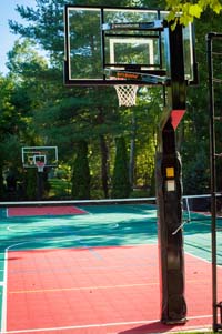 View of whole length, from hoop to hoop, of large backyard basketball court with net for tennis or volleyball, lighting system, and in-ground trampoline in Pembroke, MA.